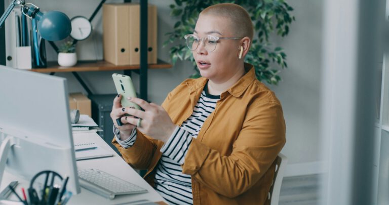 Person with a shaved head and glasses sitting at a desk, holding a smartphone and wearing wireless earbuds, appearing engaged while looking at the screen. The workspace includes a computer monitor, keyboard, notebooks, and shelves in the background.