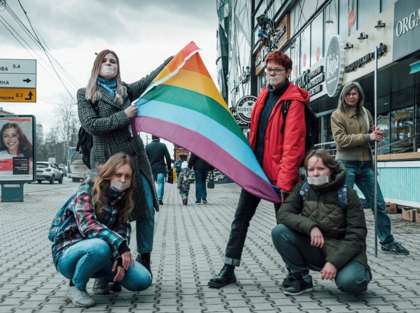 Four people with duct tape covering their mouth, two of them holding a rainbow flag