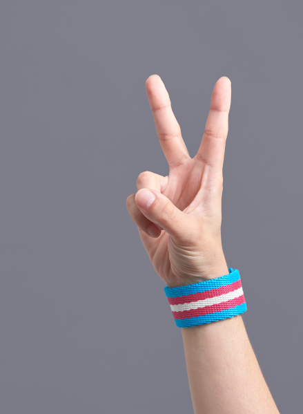 Person's hand making the V sign, wearing a bracelet featuring the colors of the trans flag.