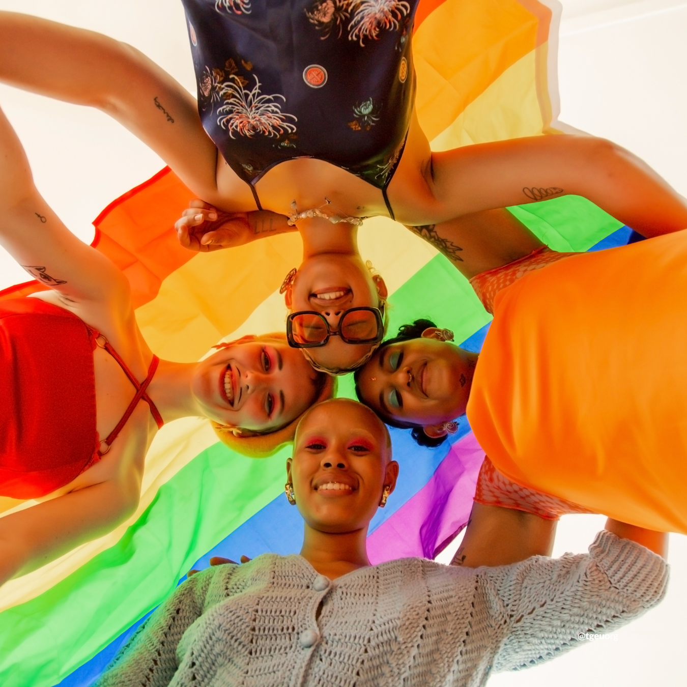 Four young people link arms and look down at the camera. A rainbow flat is draped over them.