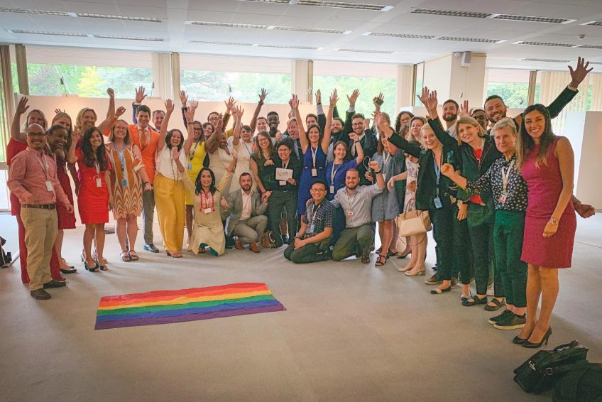 Members of the United Nations Human Rights Council posing for a group photo, waving and celebrating, with a prominent rainbow flag in the middle.