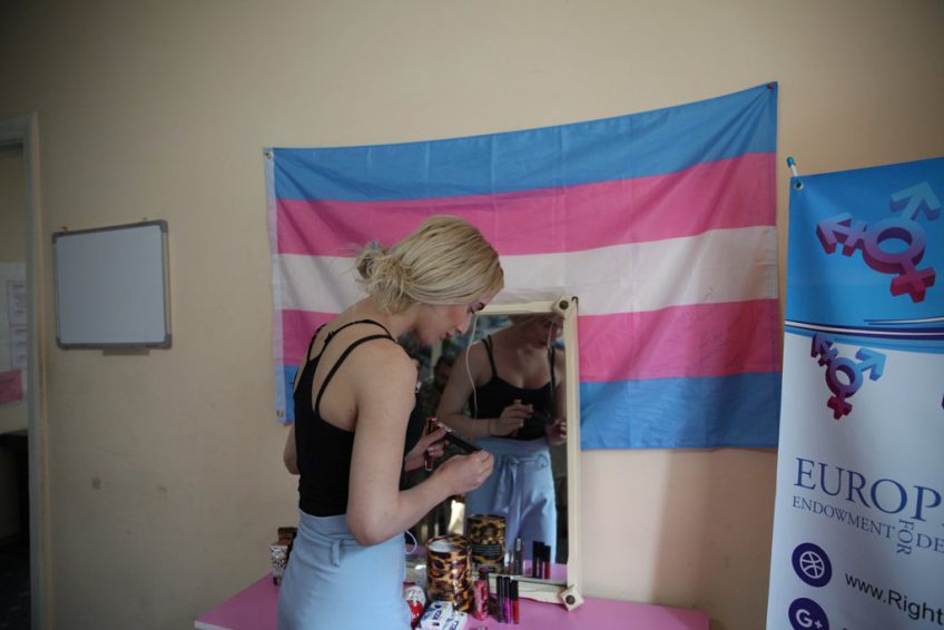 Person standing in front of a mirror with a trans flag hanging on the wall. On the right, a poster about trans rights in Europe is visible