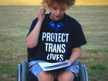 Person using a wheelchair while reading a paper. Their t-shirt says "Protect trans lives"