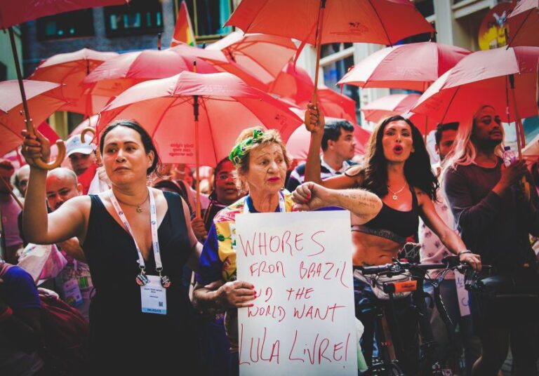 Sex workers demonstration in Amsterdam with protesters carrying red umbrellas. One of them is holding a sign that says 'Whores from Brazil and the world want Lula livre!