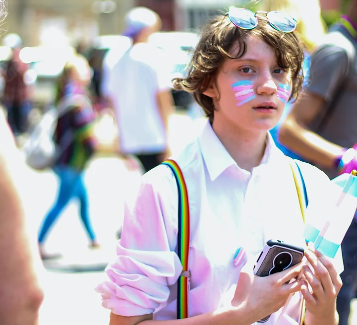 Trans youth - Young person with trans flags painted on their face, wearing rainbow suspenders and holding a little trans flag.