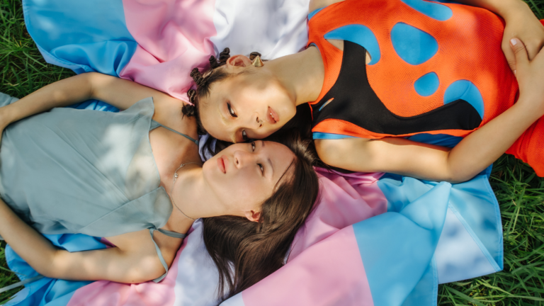 Two trans women lie on a trans pride flag.