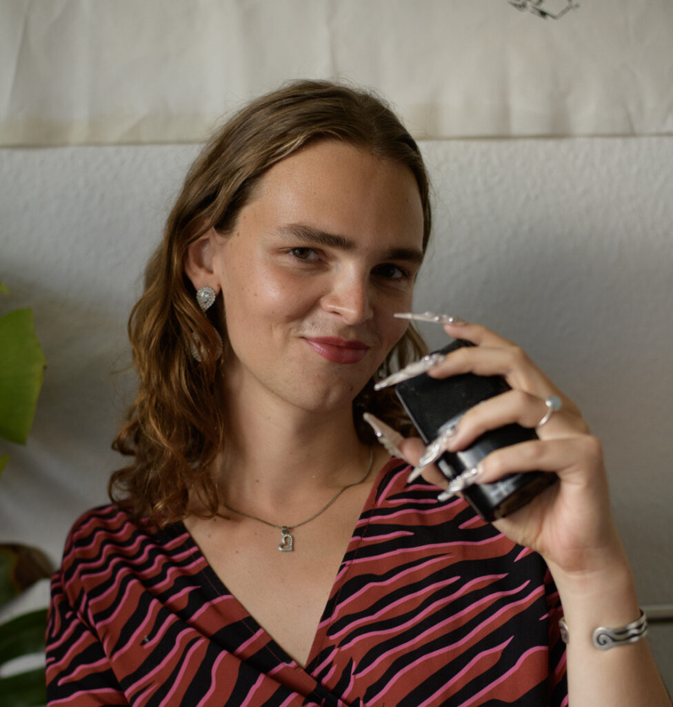 Béla, long brown hair and manicured nails holds a black microphone while smiling at the camera. She is wearing a red and black zebra-striped wrap dress, silver jewelry, and is seated indoors with a plant partially visible in the background.
