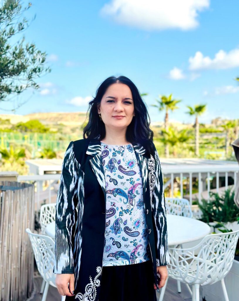 Alina (she/her), a woman with dark hair, stands outdoors on a sunny day, wearing a black and white patterned blazer over a colorful paisley-print top. She is in front of a patio area with white chairs and tables, surrounded by greenery and palm trees under a blue sky with scattered clouds.