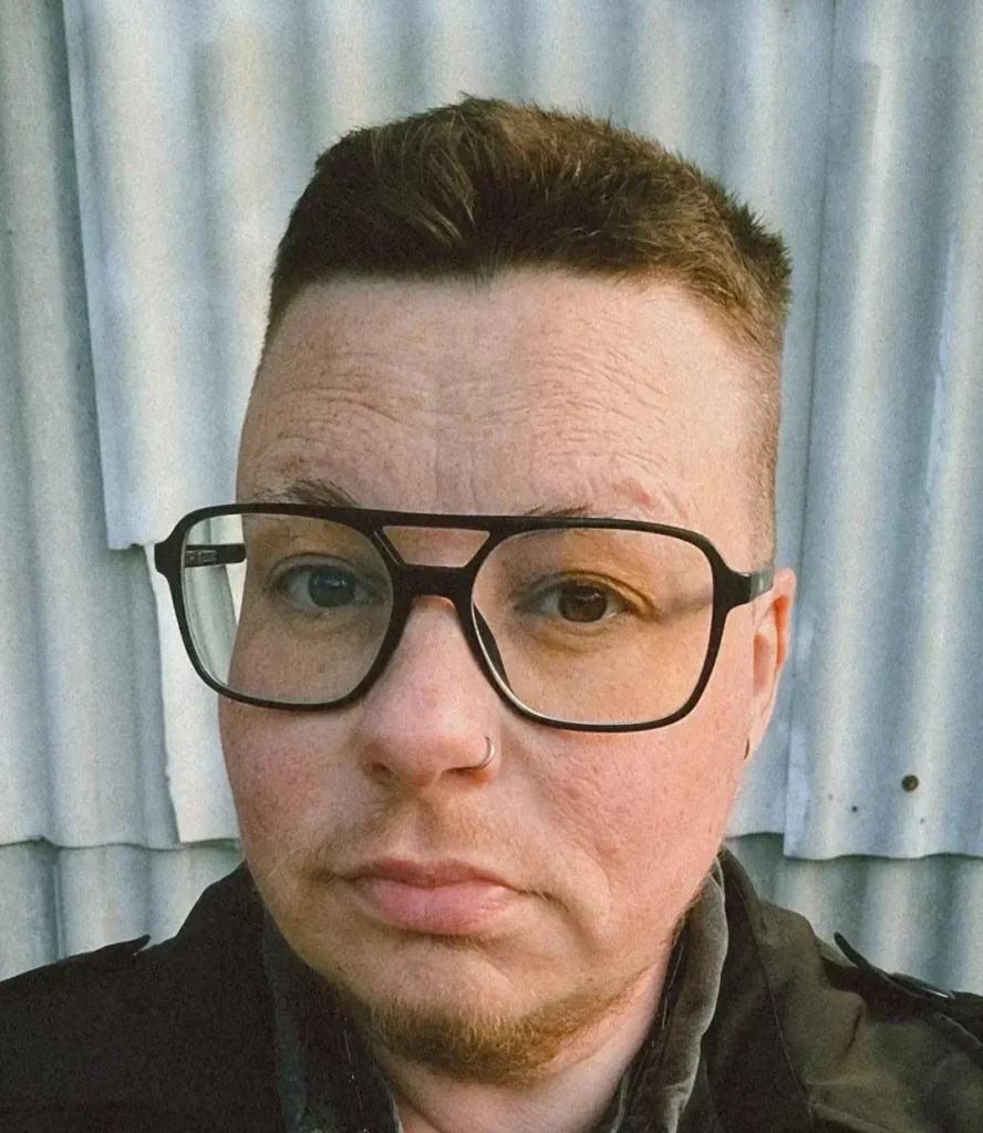 Linn (they/them) short brown hair, wearing large black-framed glasses, a nose ring, and a dark trench coat layered over a buttoned shirt, looking directly at the camera with a neutral expression.