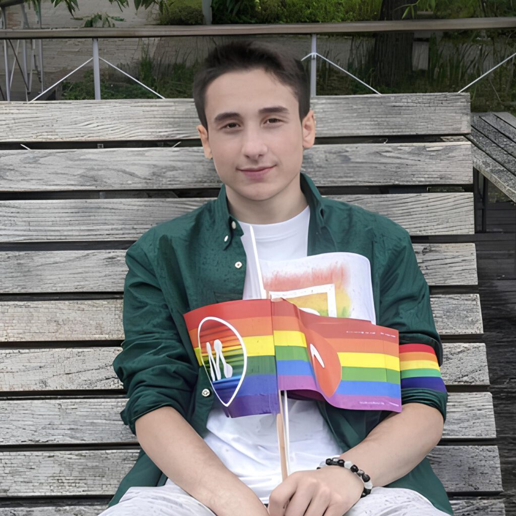 Polo with short dark hair sitting on a bench, wearing a green shirt and holding two rainbow flags