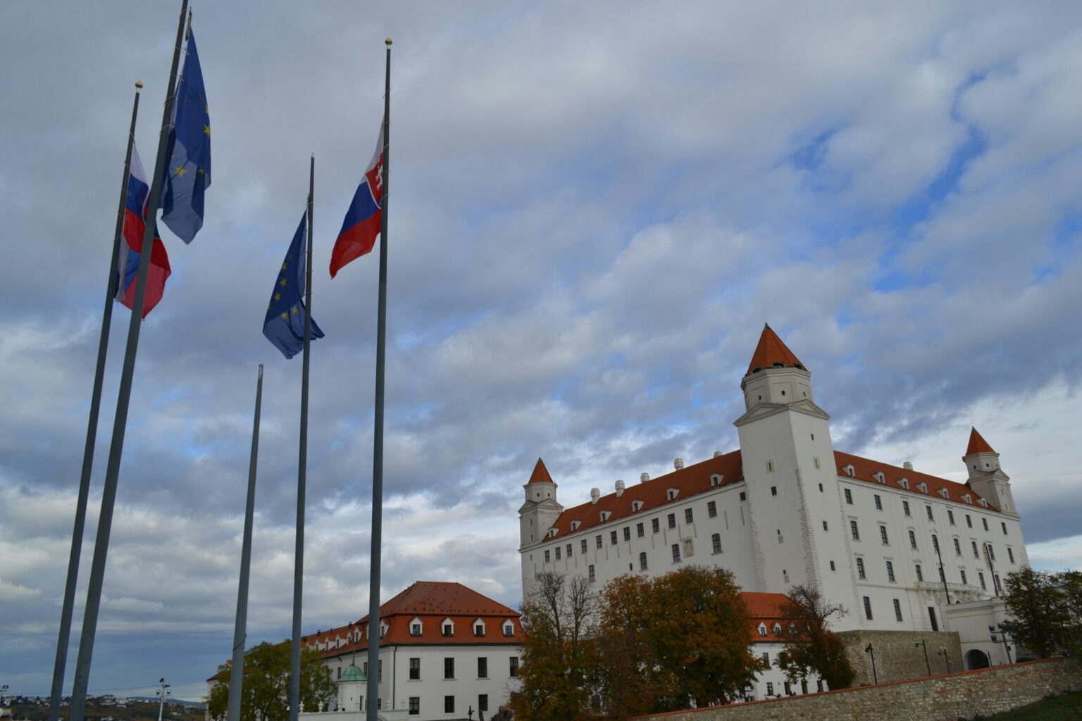 Historic castle with red-roofed towers stands under a cloudy sky. EU and Slovakia flag flutter nearby, adding vibrancy to the scene.