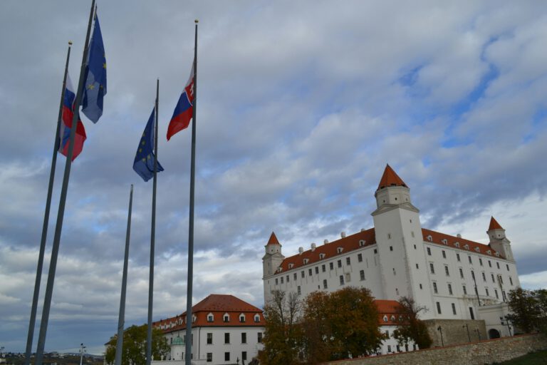 Historic castle with red-roofed towers stands under a cloudy sky. EU and Slovakia flag flutter nearby, adding vibrancy to the scene.