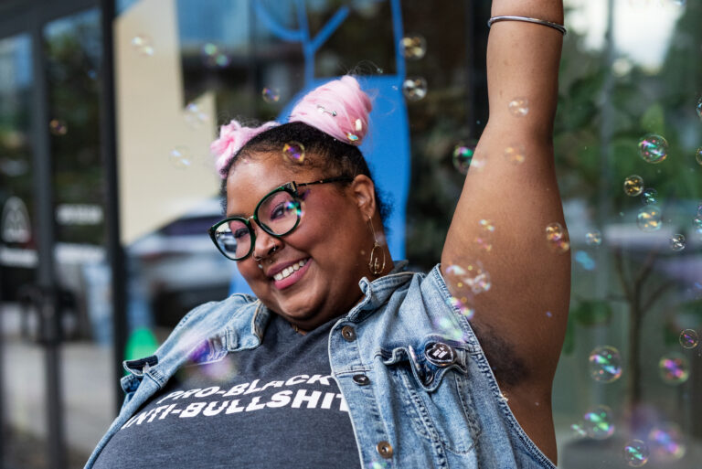 A non-binary Black person with glasses and space buns. They’re modeling a tank top reading “Pro-Black” on one line and “Anti-Bullshit” below, complemented by a denim vest.