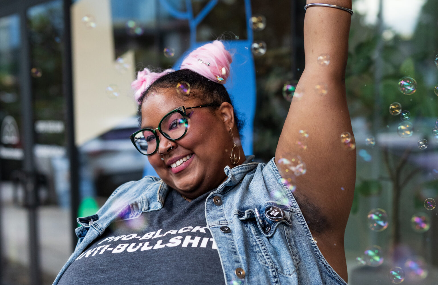 A non-binary Black person with tinted sunglasses and space buns leans back in a chair, their face mask adorned with a gold double chain. They’re modeling a tank top reading “Pro-Black” on one line and “Anti-Bullshit” below, complemented by a denim vest.